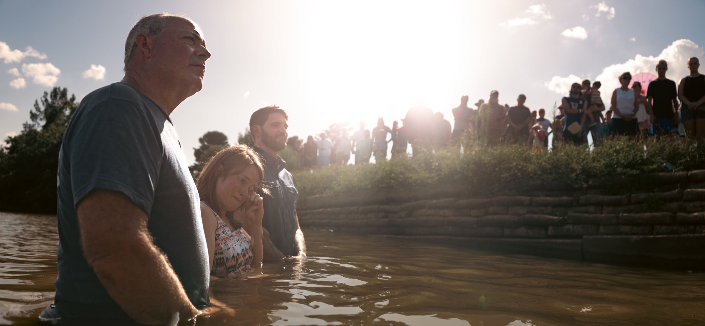 A baptism in a river, the congregation watching from the bank against a bright sky.