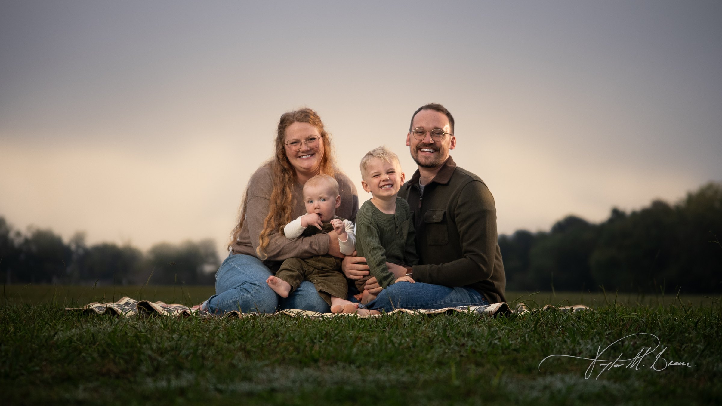The Gibson family, two parents and two little boys, smiling together on a blanket in an open field at dusk.