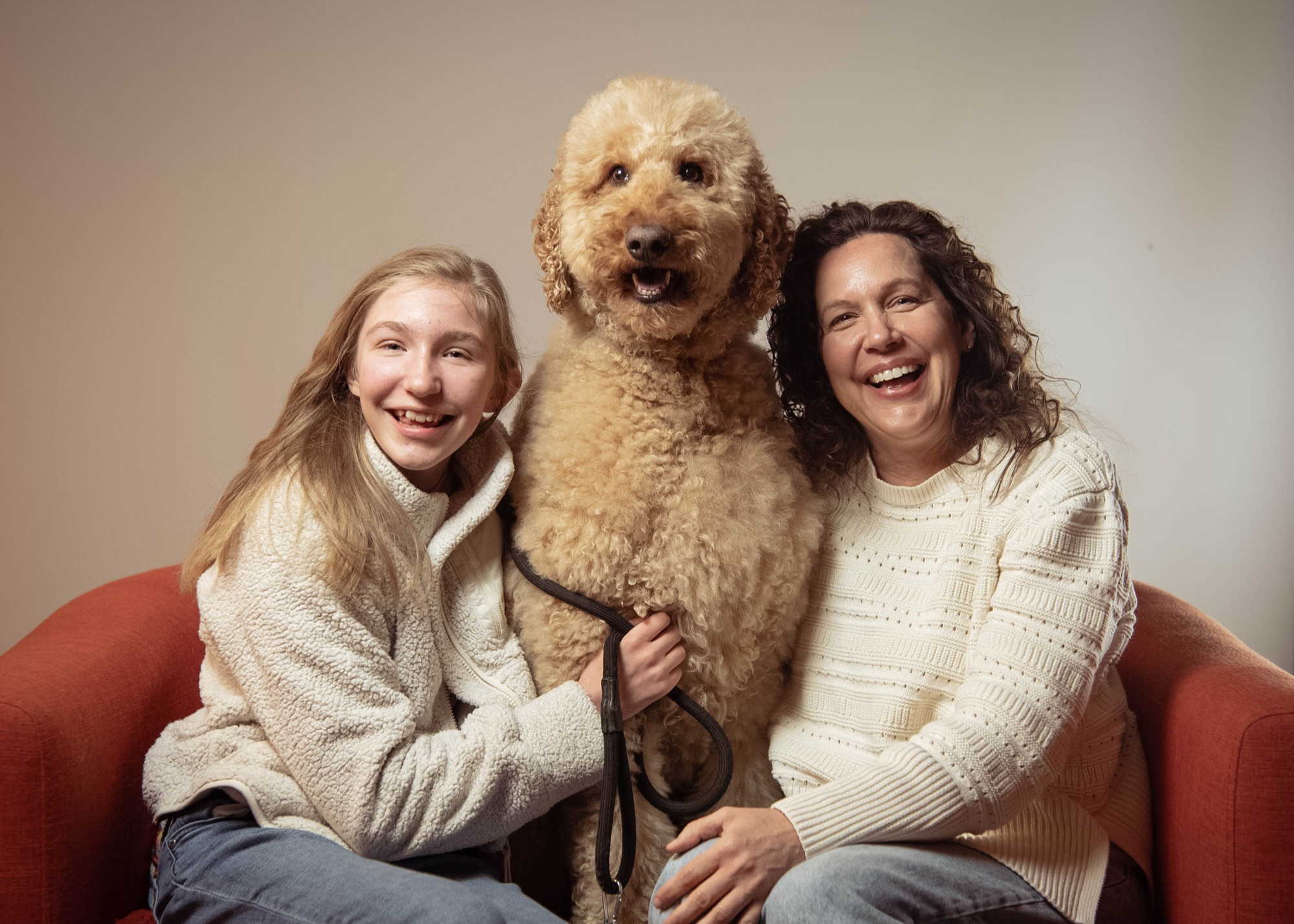 A young woman and her mother laughing on a red couch with their big curly dog between them.