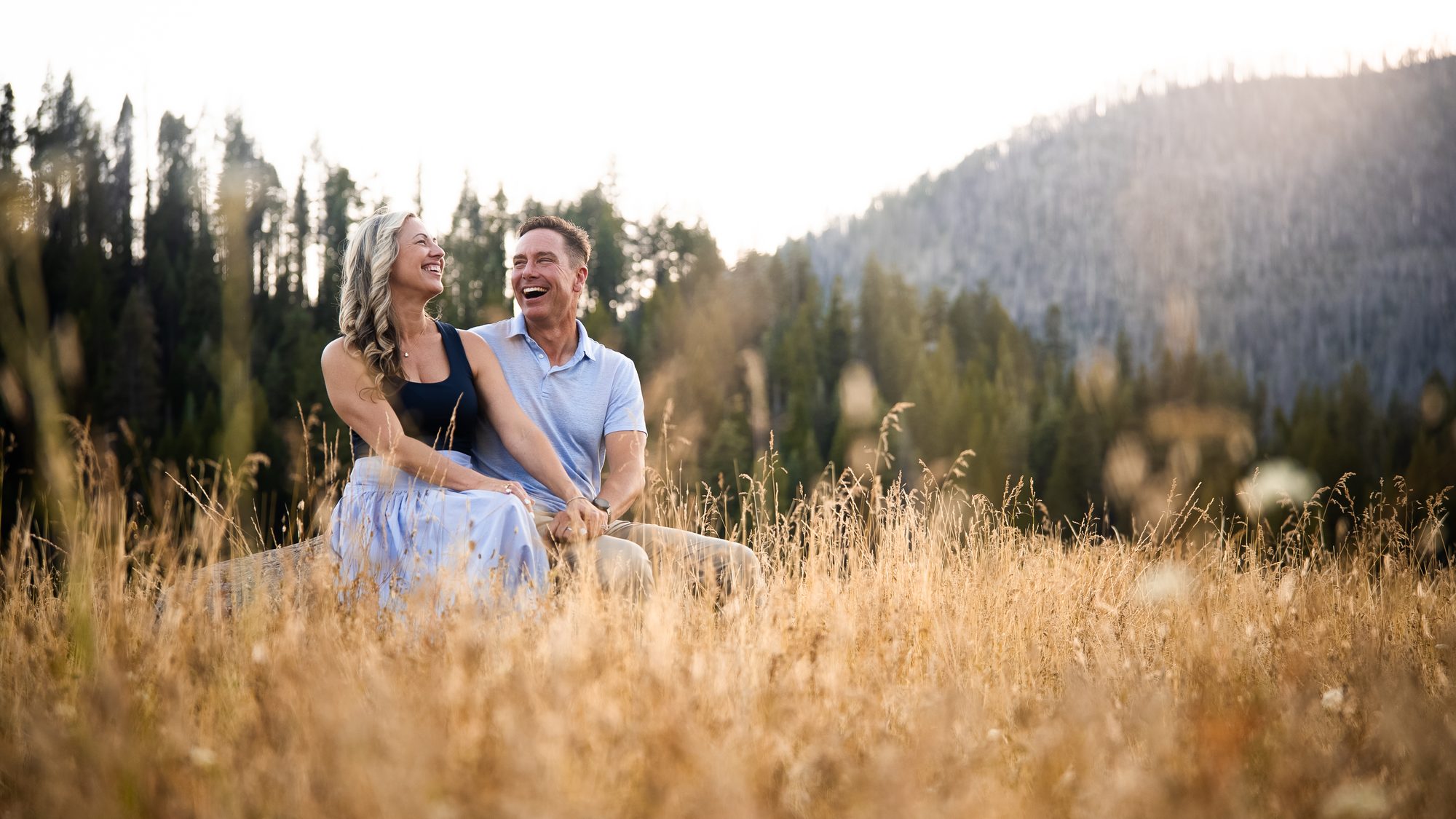 A couple sitting together in a golden meadow below pine-covered hills.