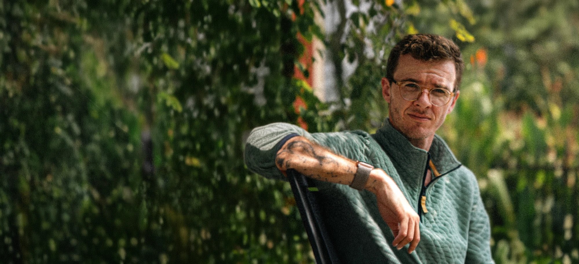 Taylor Mac Brown, seated, arm resting on the back of a chair, in front of green leaves.